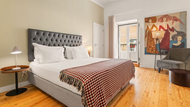 A bedroom with a tufted headboard, a large bed with white linens, a red and black checkered throw blanket, a grey sofa, a floor lamp, and a painting on the wall. - Casa do Bairro by Shiadu (Lisbon)