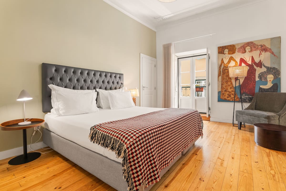 A bedroom with a tufted headboard, a large bed with white linens, a red and black checkered throw blanket, a grey sofa, a floor lamp, and a painting on the wall.