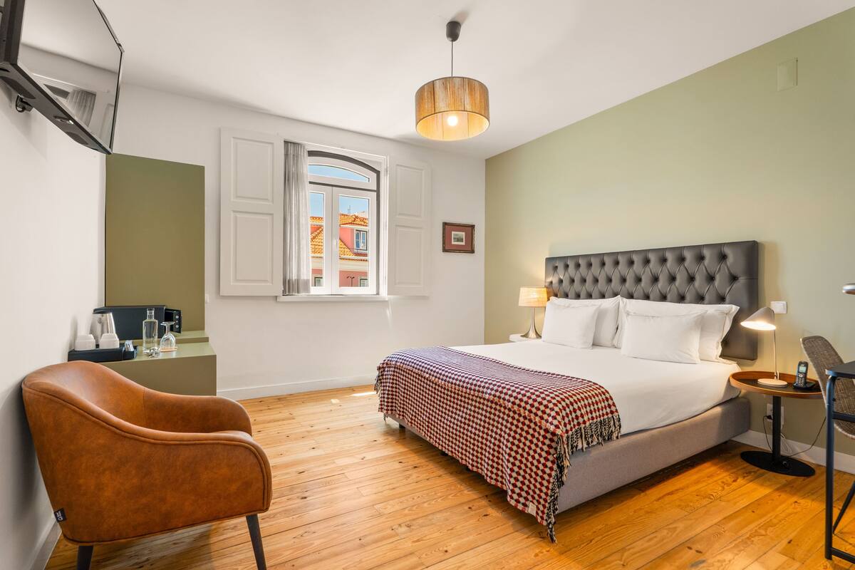 A hotel room with a large bed, a wooden floor, a chair, a desk with a lamp, and a window with curtains.