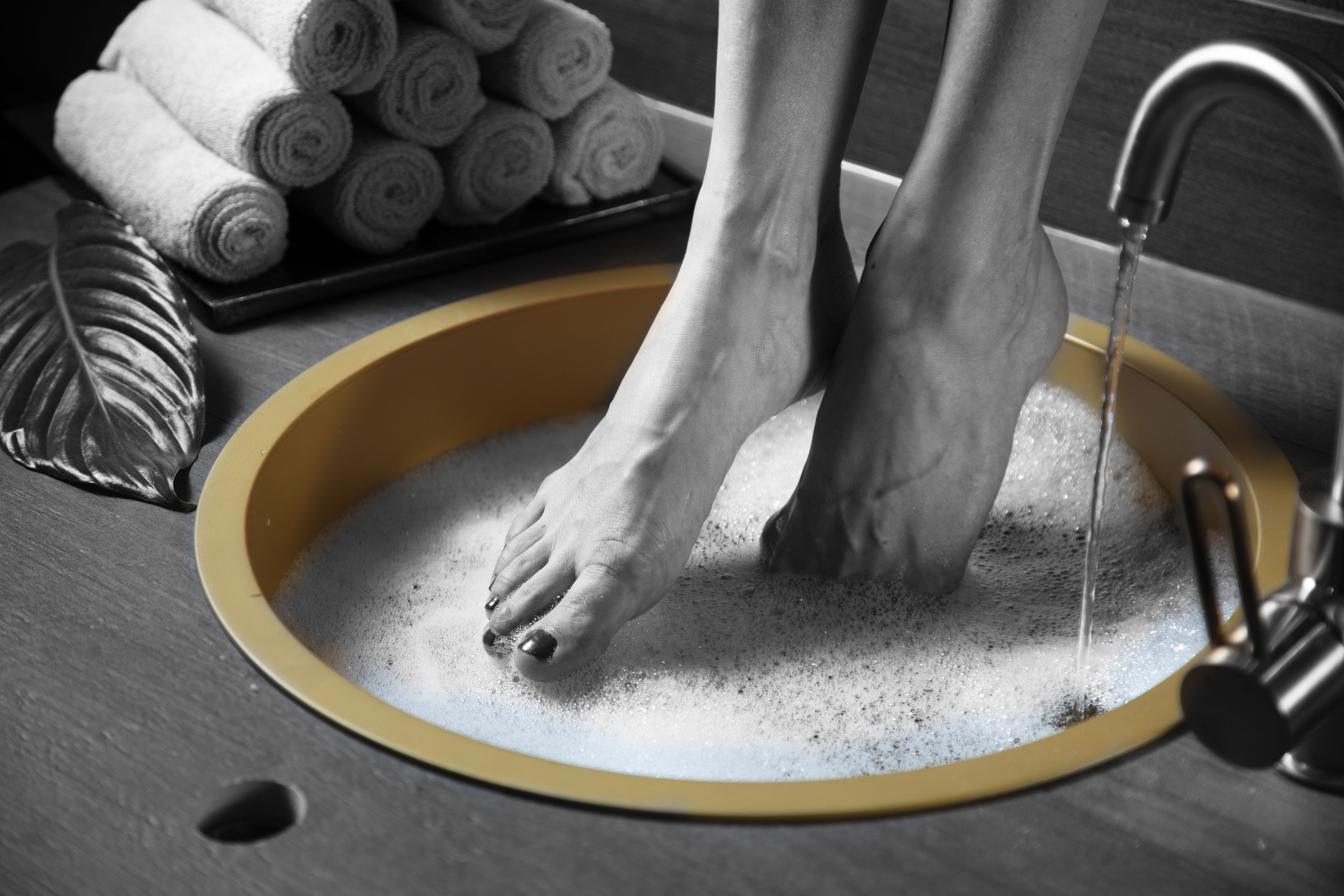 Un lavamanos con borde amarillo, una persona lavándose los pies en él y una bandeja con toallas enrolladas al fondo.