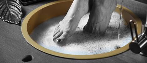 Un lavamanos con borde amarillo, una persona lavándose los pies en él y una bandeja con toallas enrolladas al fondo.