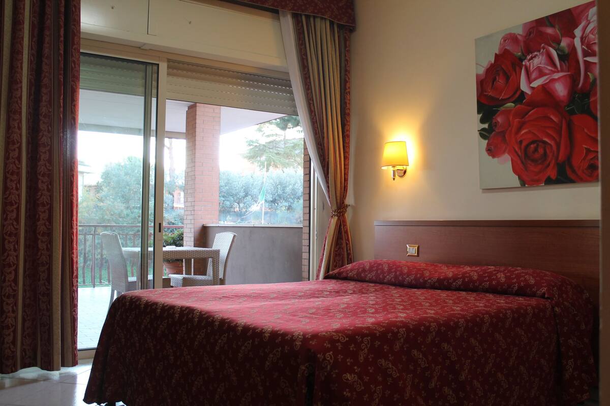 A hotel room with a bed, a balcony with a table and chairs, and a painting of roses on the wall.