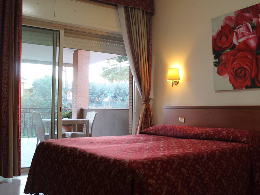 Una habitación de hotel con cama, un balcón con mesa y sillas, y un cuadro de rosas en la pared.