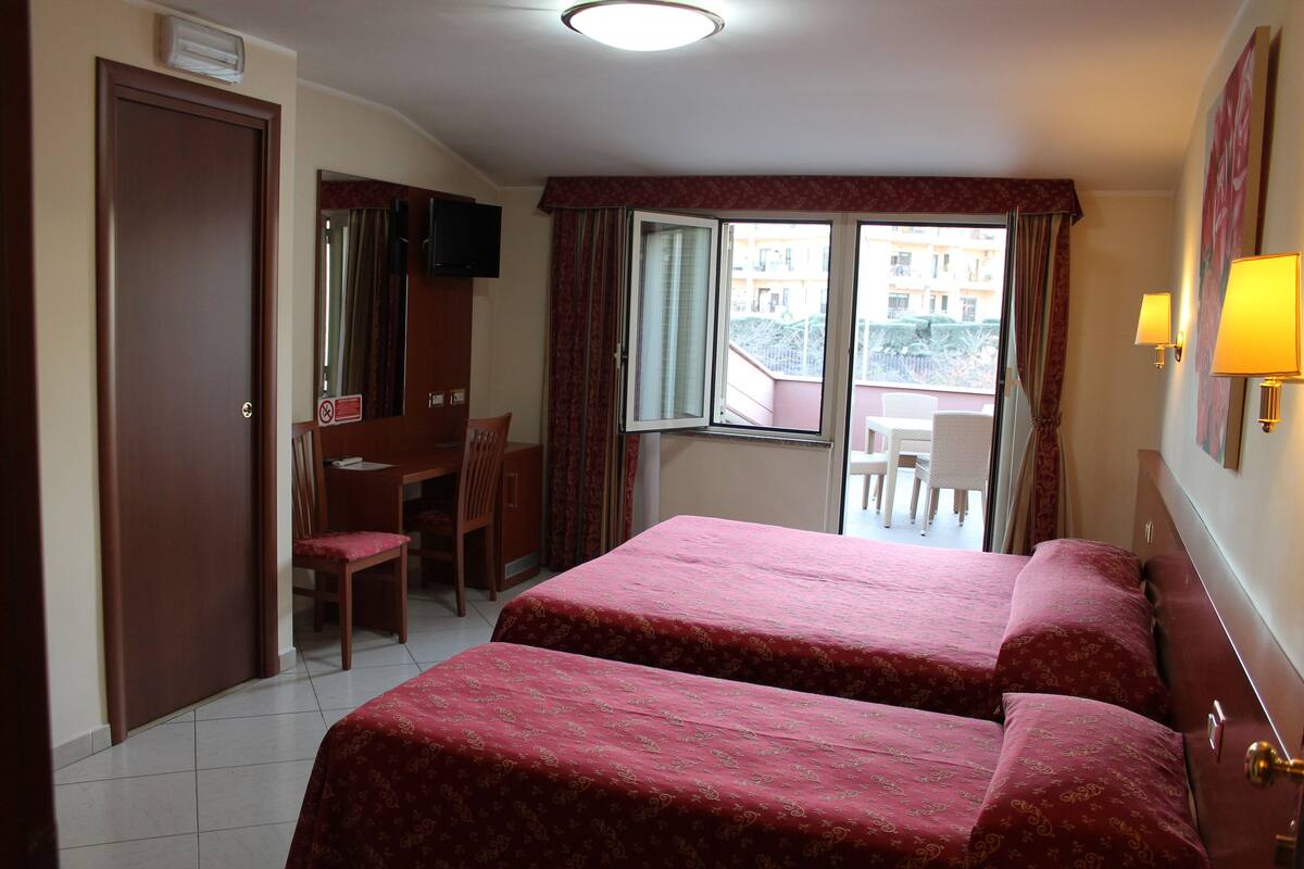 A hotel room with two beds, a desk, a chair, a television, and a balcony with a view of buildings.