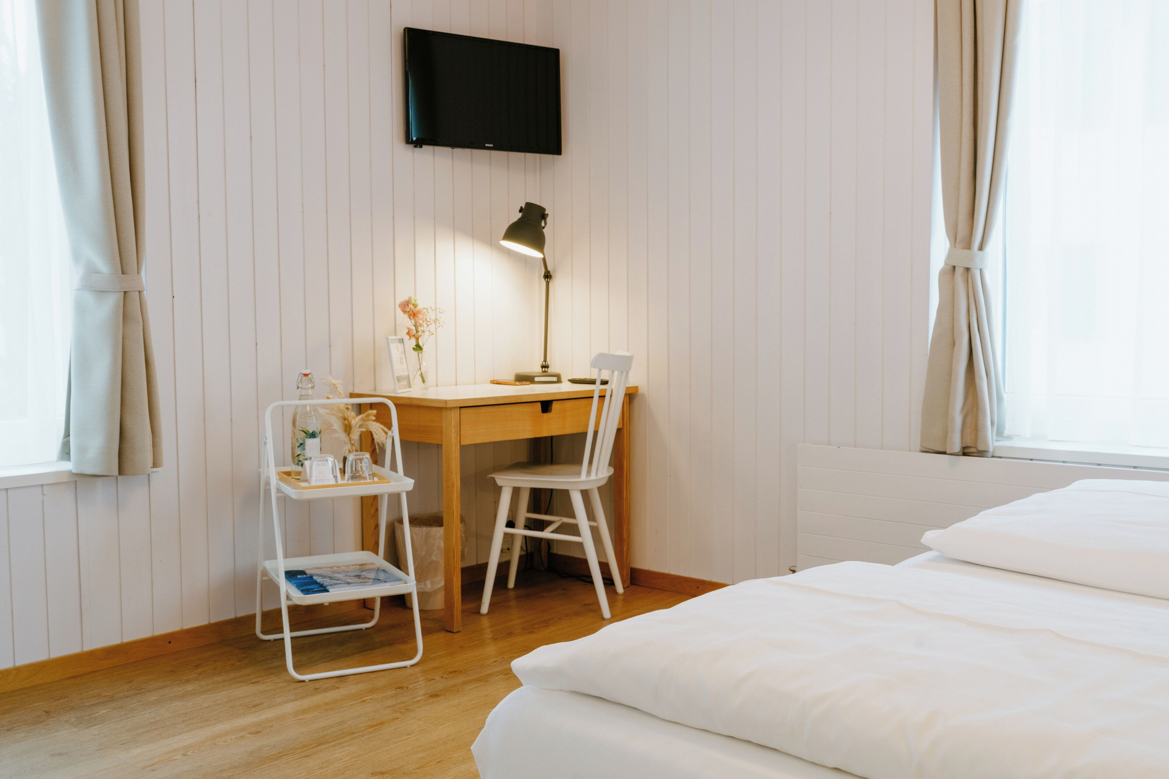 A bedroom with a wooden desk, a chair, a bed, and a TV mounted on the wall.