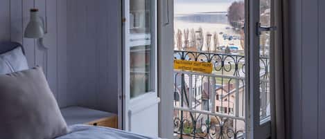 Un dormitorio con una cama, una mesita de noche, una lámpara y vistas a un lago y montañas a través de una puerta abierta.