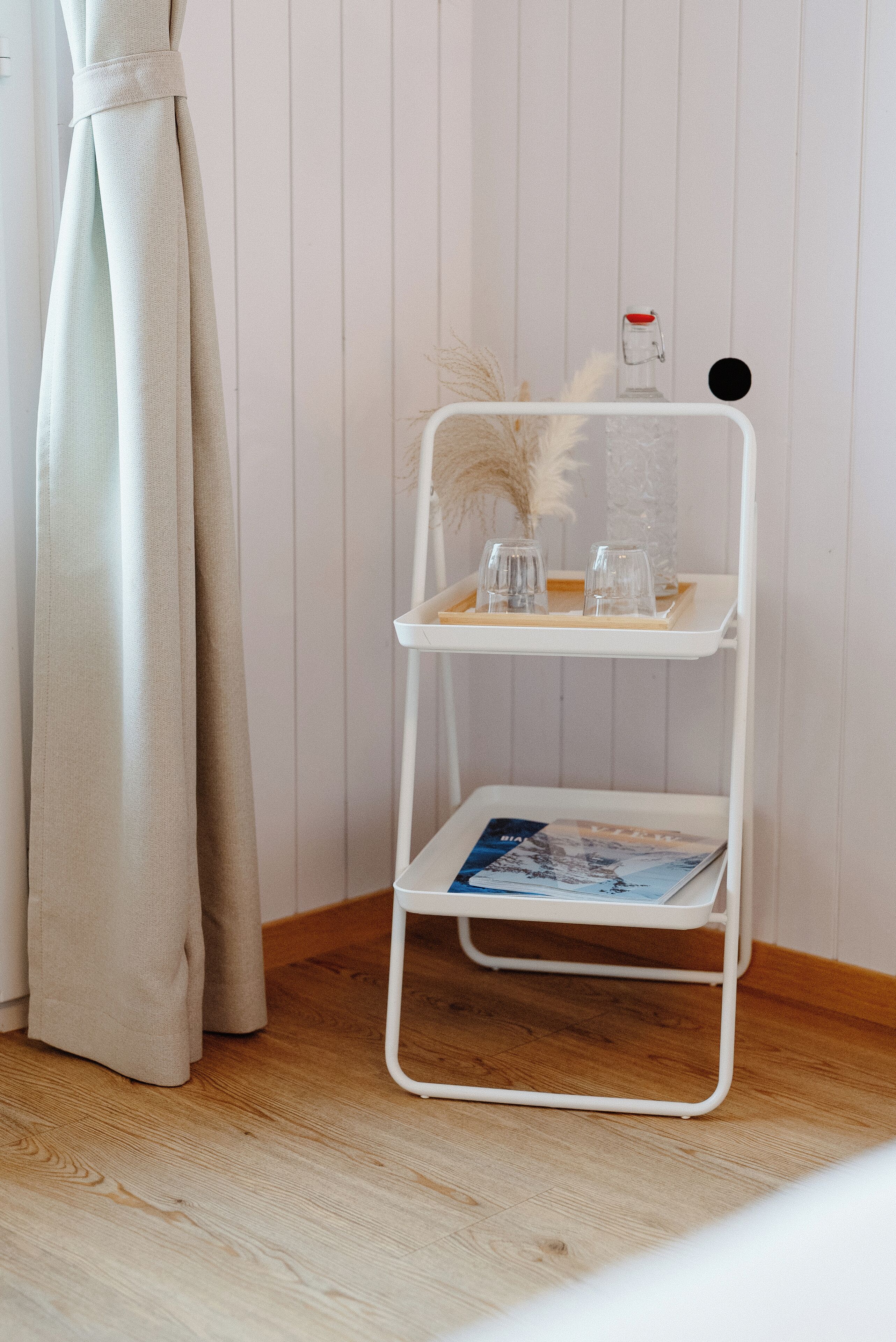 A white metal shelf with glassware and a magazine, placed against a wooden floor and white wall.