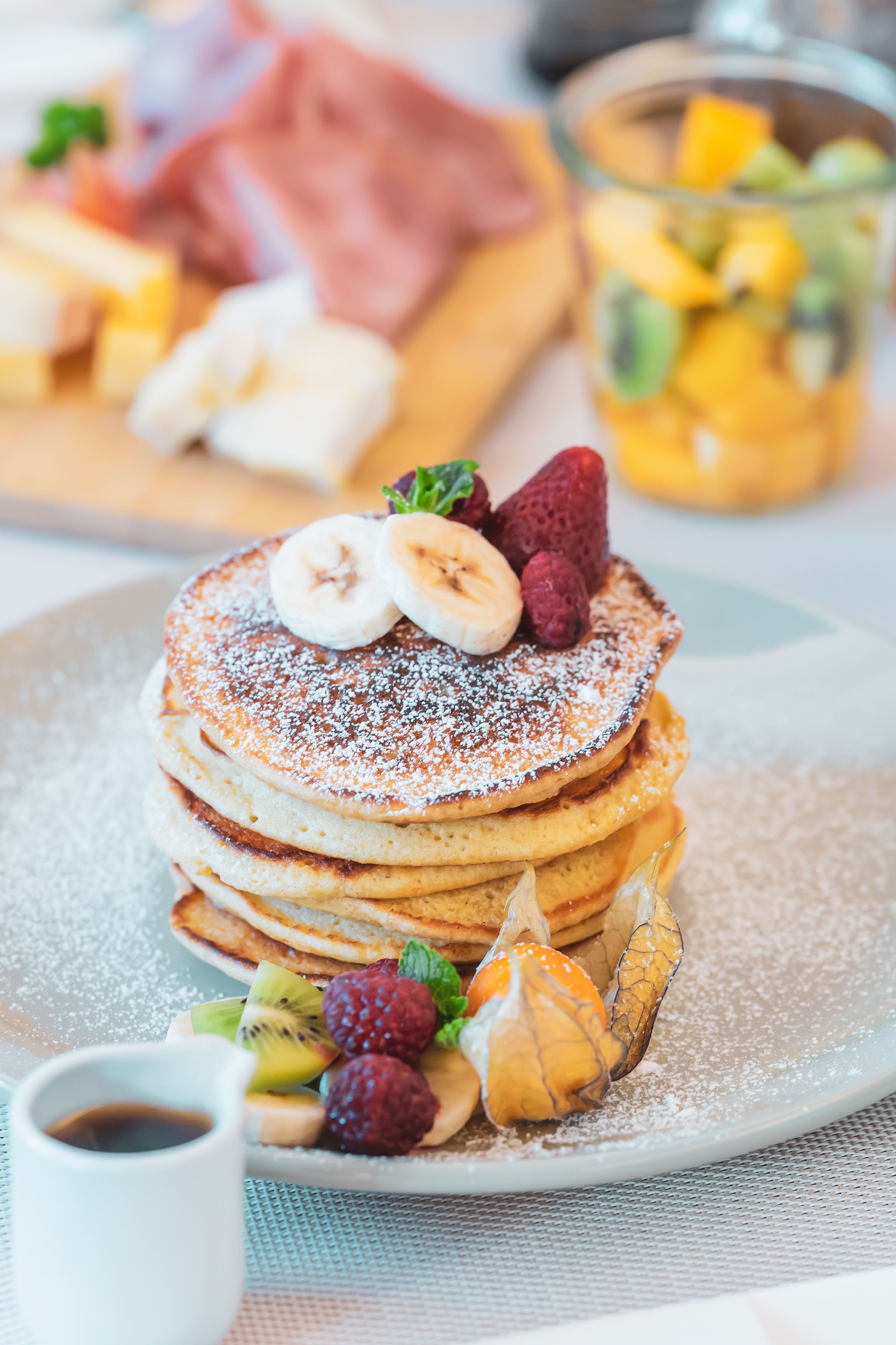 A stack of pancakes with fruit toppings and powdered sugar, accompanied by a small cup of syrup and a glass of fruit salad.