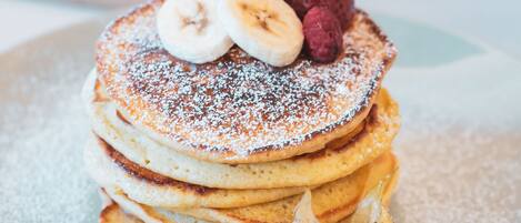 A stack of pancakes with fruit toppings and powdered sugar, accompanied by a small cup of syrup and a glass of fruit salad.