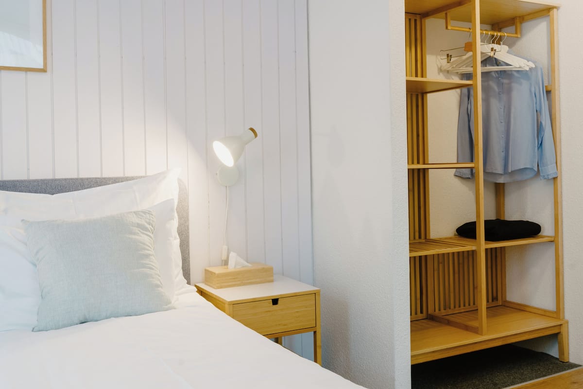A neatly made bed with white linens, a bedside table with a lamp, and a wooden wardrobe with hanging clothes.