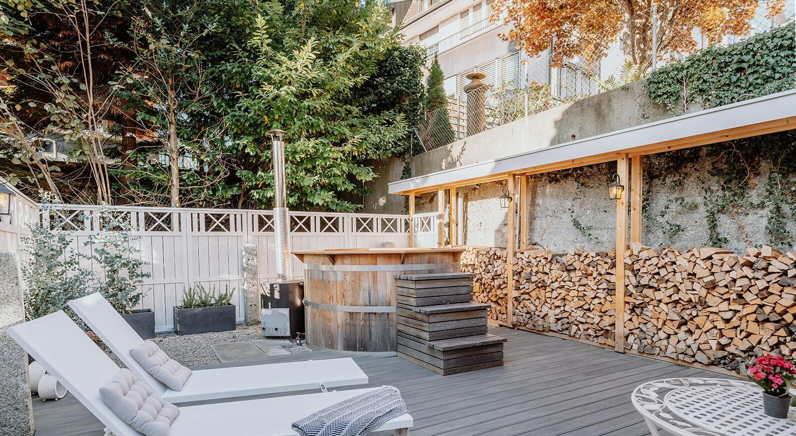 Terraza de madera con sillones de descanso, zona de barbacoa y leña apilada.