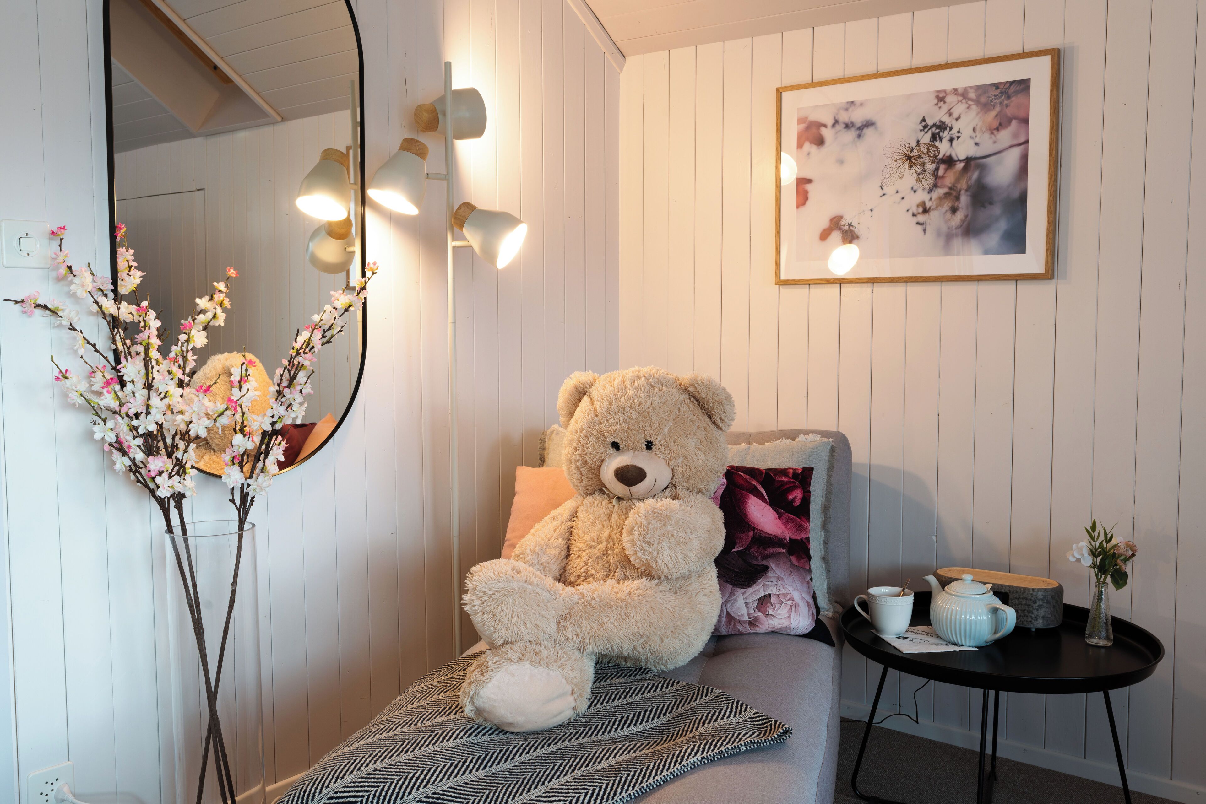Una habitación con un gran osito de peluche sentado en un sofá, un espejo, un jarrón con flores, una mesita con una tetera y un cuadro en la pared.