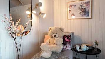 Una habitación con un gran osito de peluche sentado en un sofá, un espejo, un jarrón con flores, una mesita con una tetera y un cuadro en la pared.