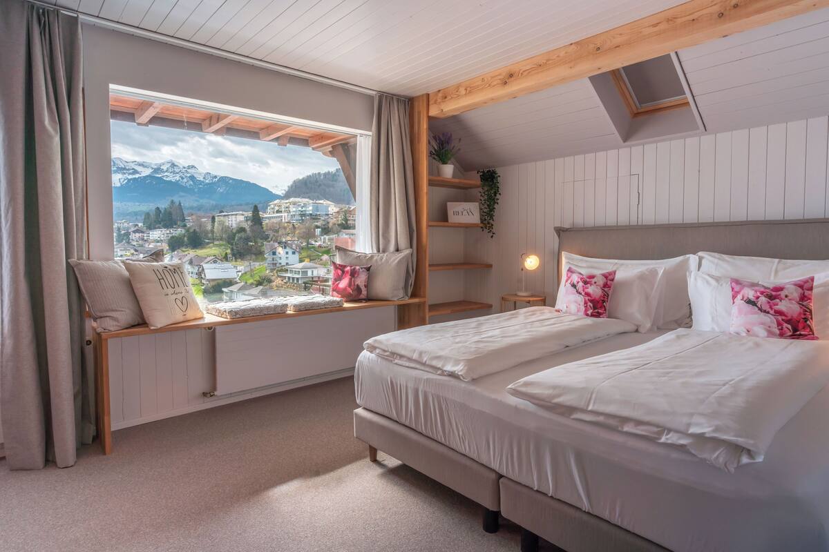 A bedroom with a large window, a bed, a bedside table, and a shelf with a plant.