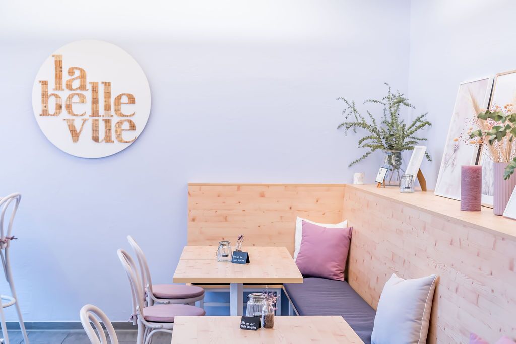 A cozy cafe interior with a wooden bench, two tables, and chairs. A round sign with "la belle ville" is on the wall.