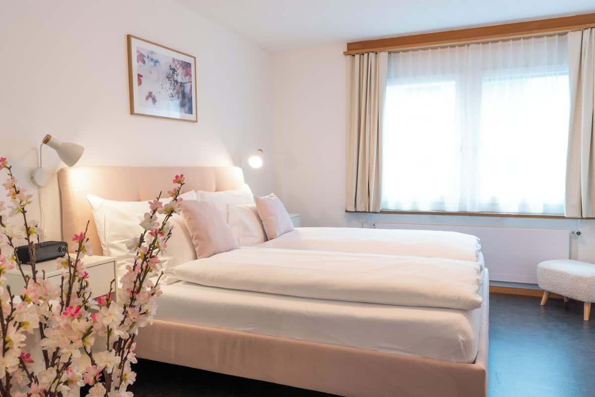 A neatly arranged bedroom with a large bed, bedside lamps, a small table, and a window with curtains.