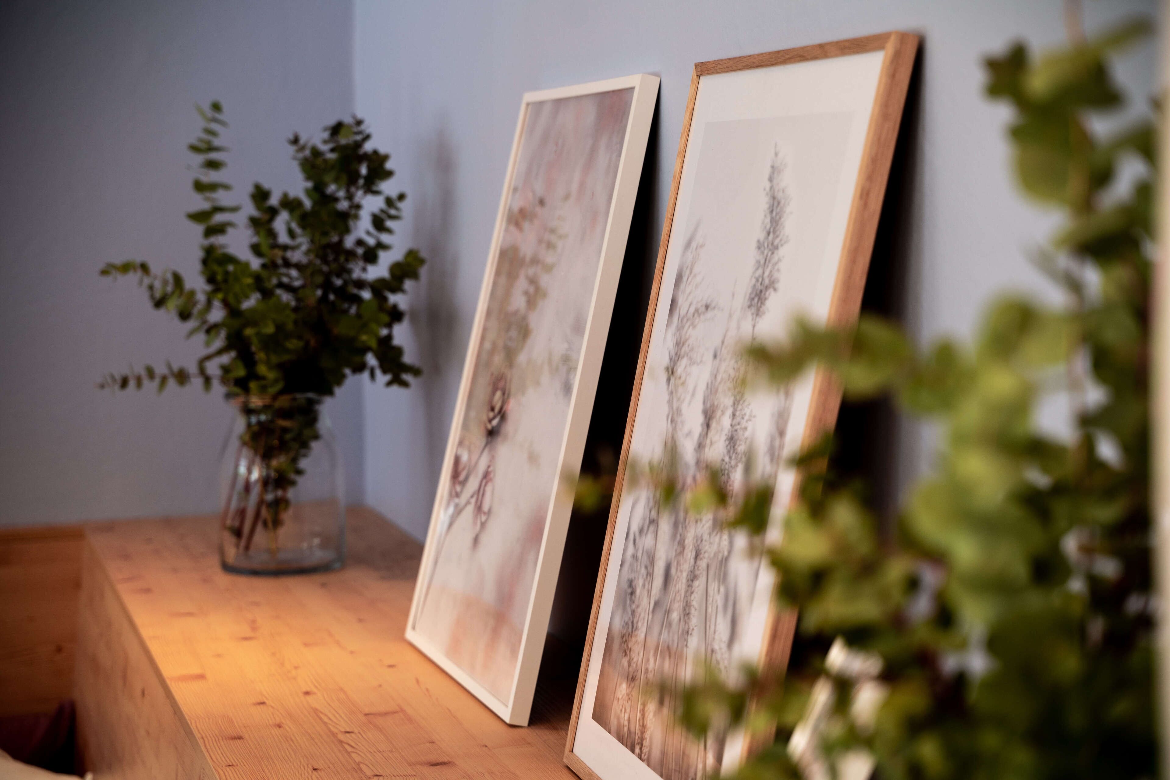 Deux œuvres d’art encadrées sont posées sur une surface en bois, avec un vase de plantes vertes en arrière-plan.