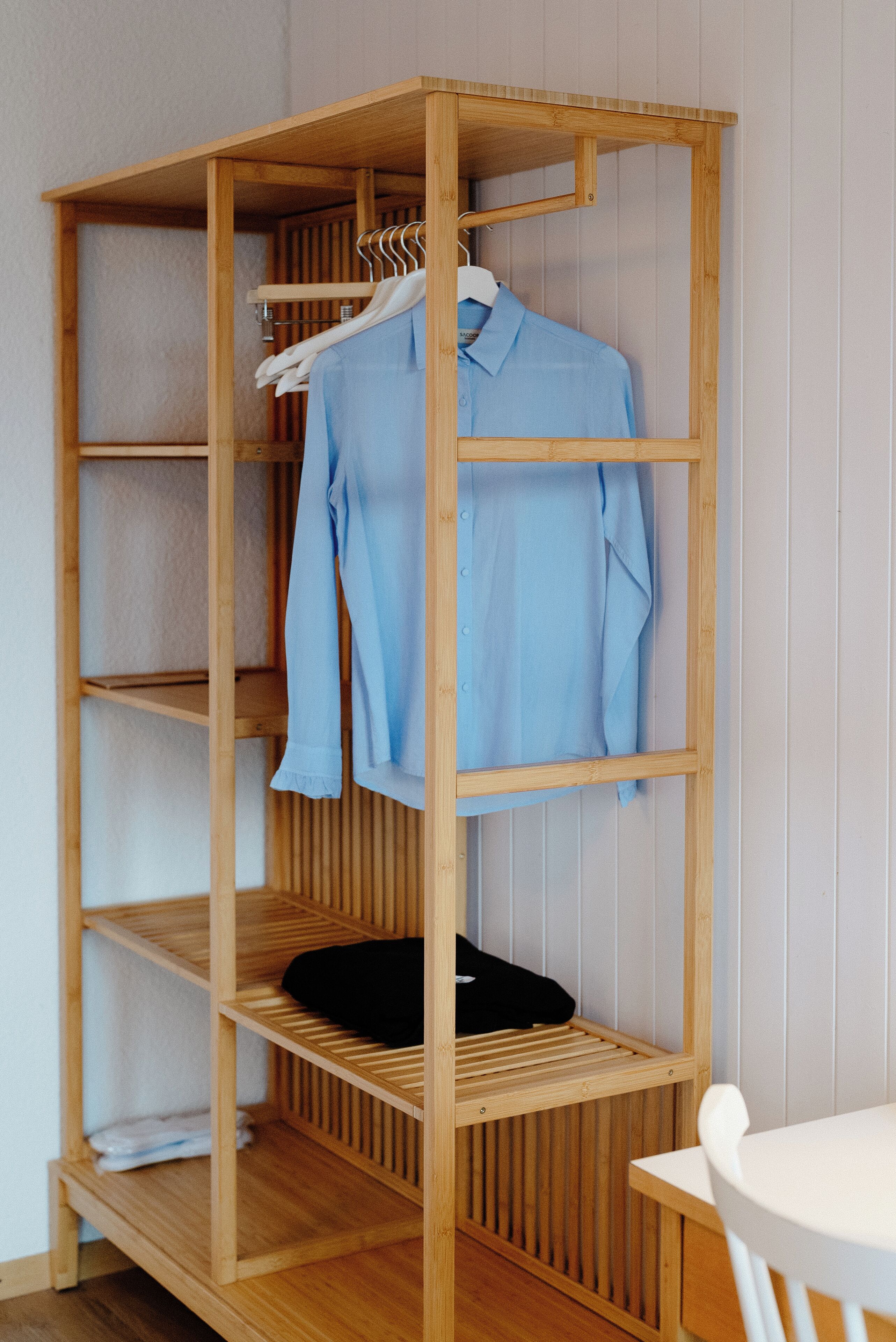 Ein Holzschrank mit mehreren Einlegeböden und einer Kleiderstange, gefüllt mit gefalteter Kleidung und einem aufgehängten Hemd.