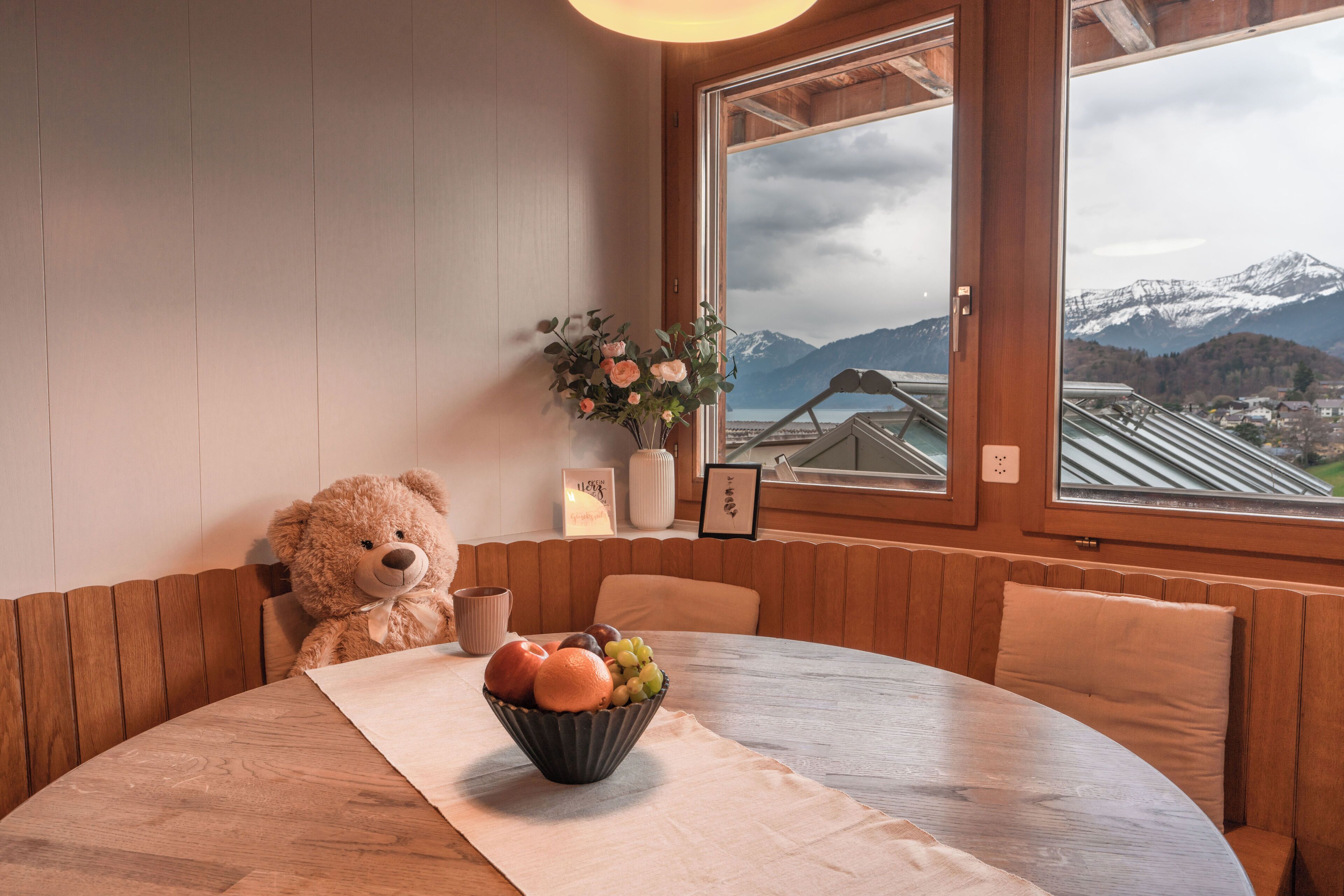 A cozy dining area with a wooden table, a bowl of fruit, a teddy bear, and a view of mountains through a window.