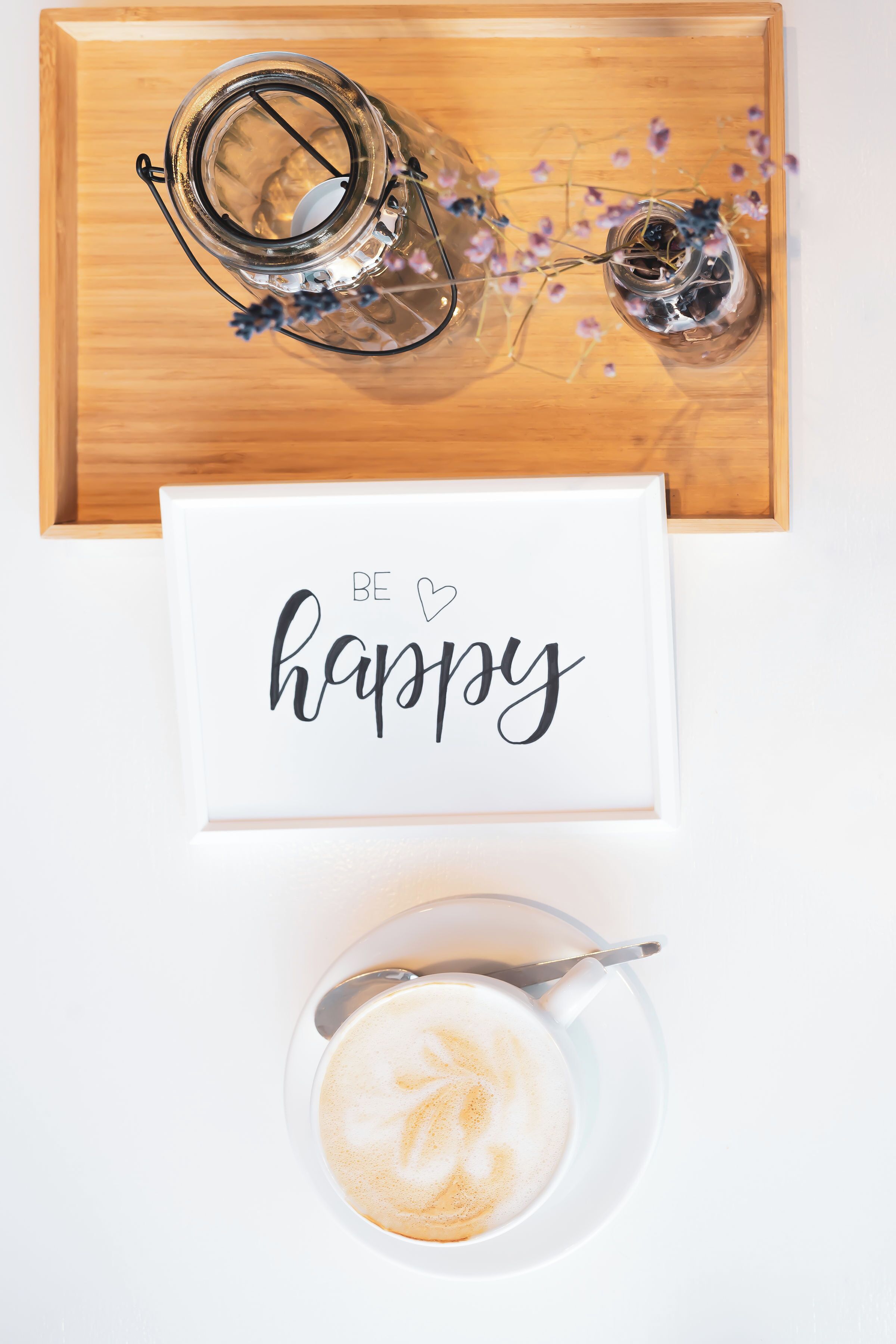 A wooden tray with two glass jars containing dried flowers, a framed sign with the words "BE happy", and a cup of coffee with latte art.