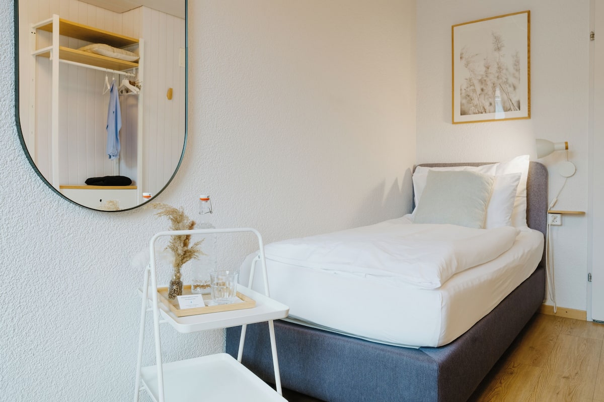 A bedroom with a bed, a bedside table with a tray, a mirror, a wardrobe, and a framed picture on the wall.
