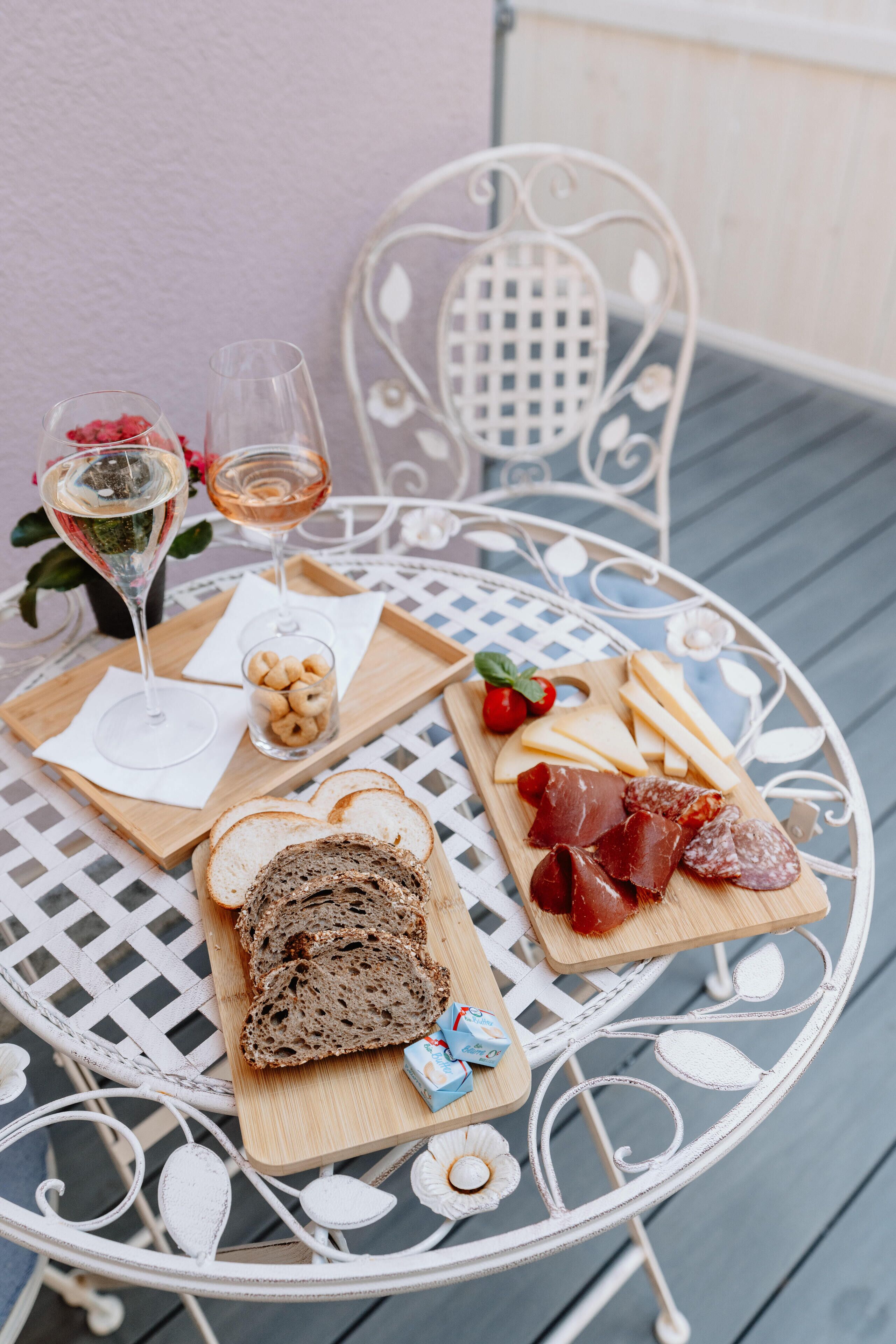 Una mesa metálica redonda con un diseño de celosía blanca, presentando una tabla de madera con pan, queso y embutidos, dos copas de vino rosado y un tazón pequeño de frutos secos.
