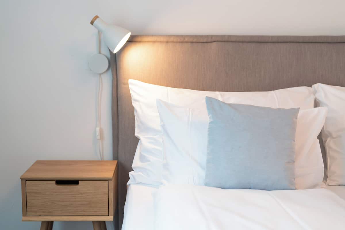 A neatly made bed with white and light blue pillows, a wooden bedside table, and a wall-mounted lamp.