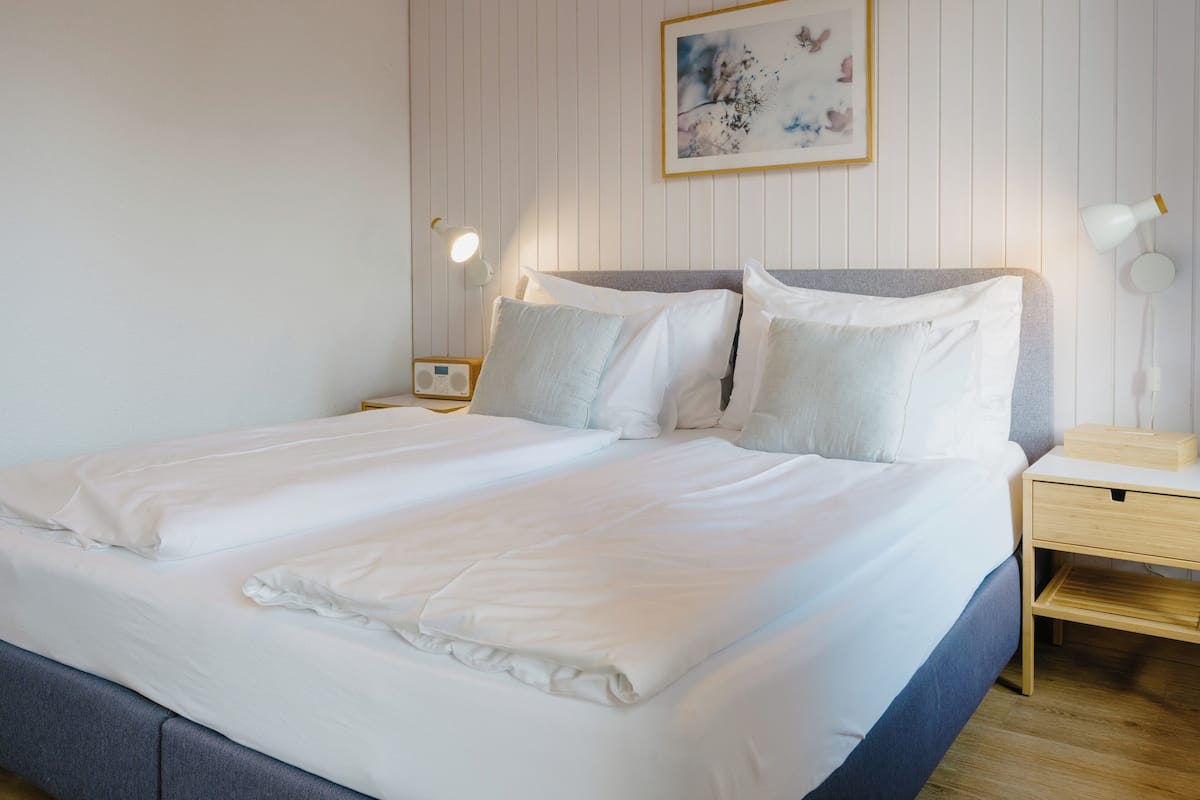 A neatly made bed with white linens, a bedside table with a lamp, and a wall-mounted picture frame.