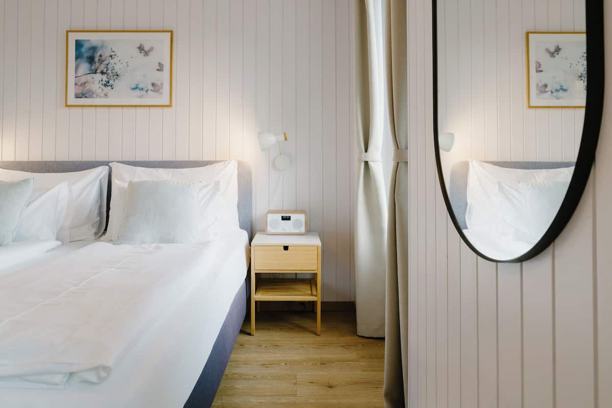 A neatly made bed with white linens, a bedside table with a radio, and a mirror on the wall.