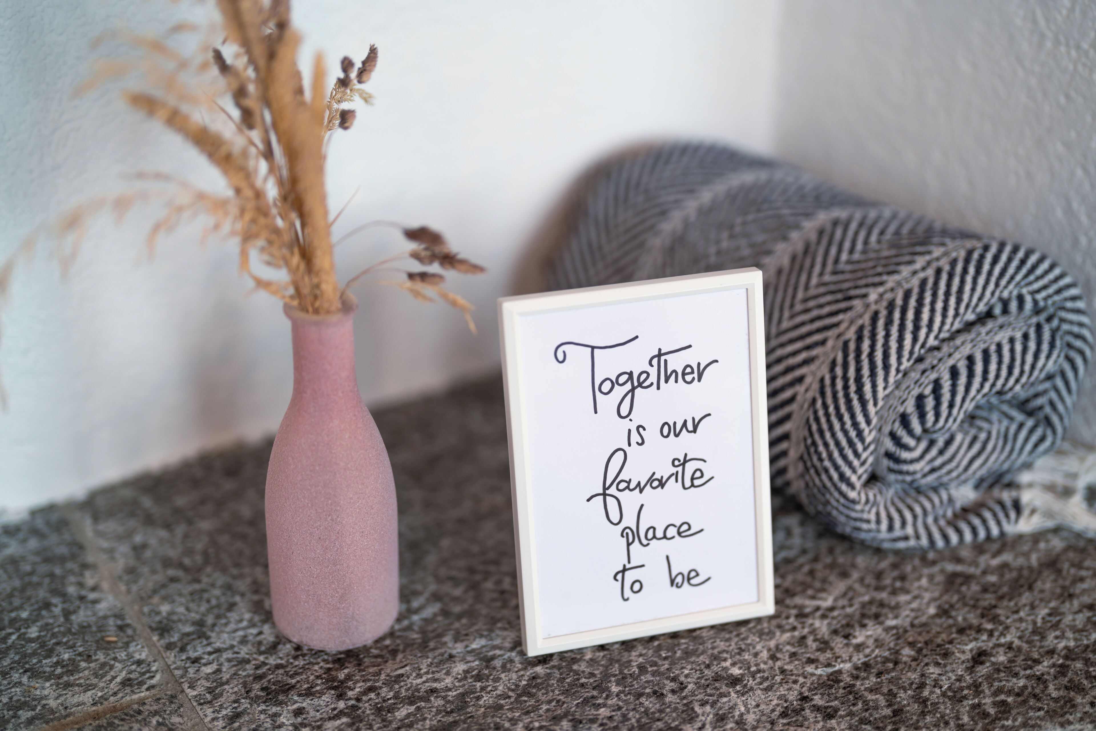 Una frase inspiradora enmarcada, un jarrón rosa con plantas secas y una manta de punto gris sobre una superficie de piedra.