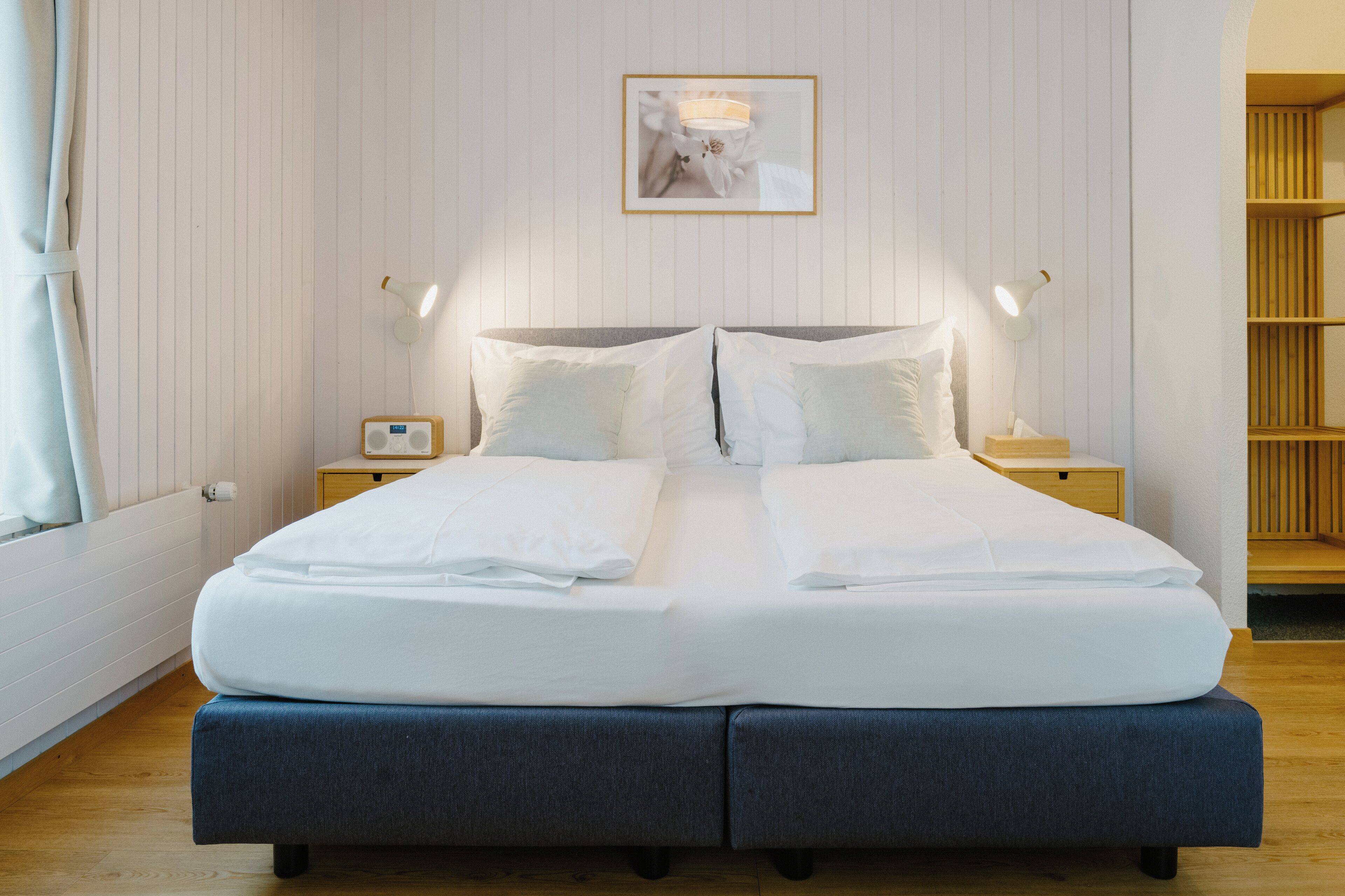 A neatly made double bed with white linens, two bedside tables with lamps, a framed picture on the wall, and a wooden bookshelf.