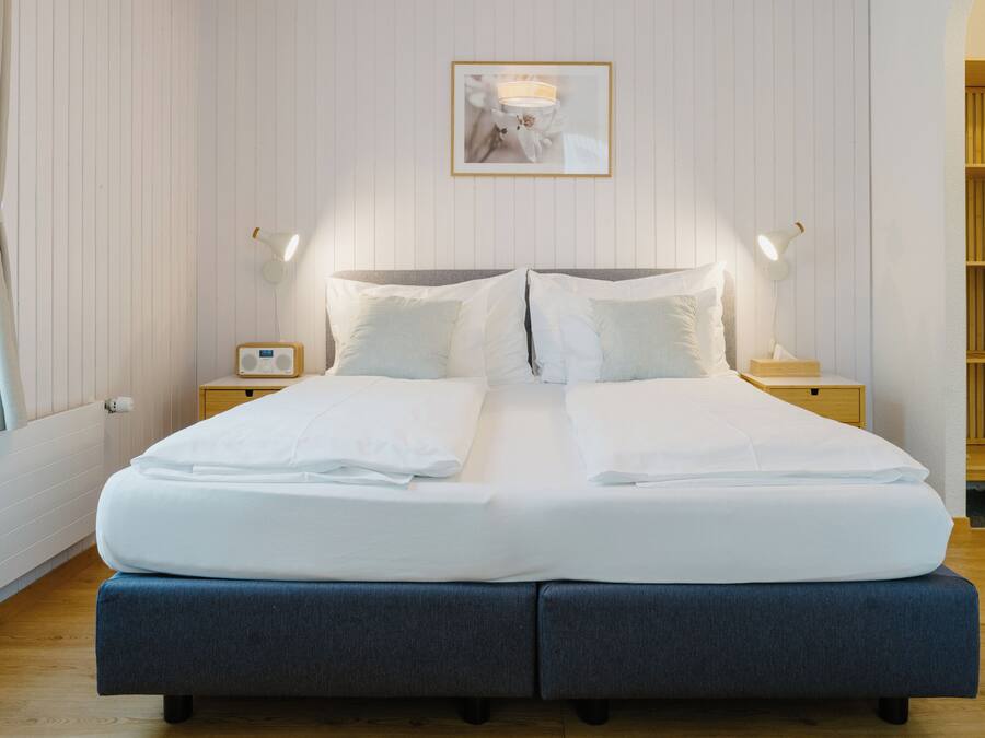 A neatly made double bed with white linens, two bedside tables with lamps, a framed picture on the wall, and a wooden bookshelf.
