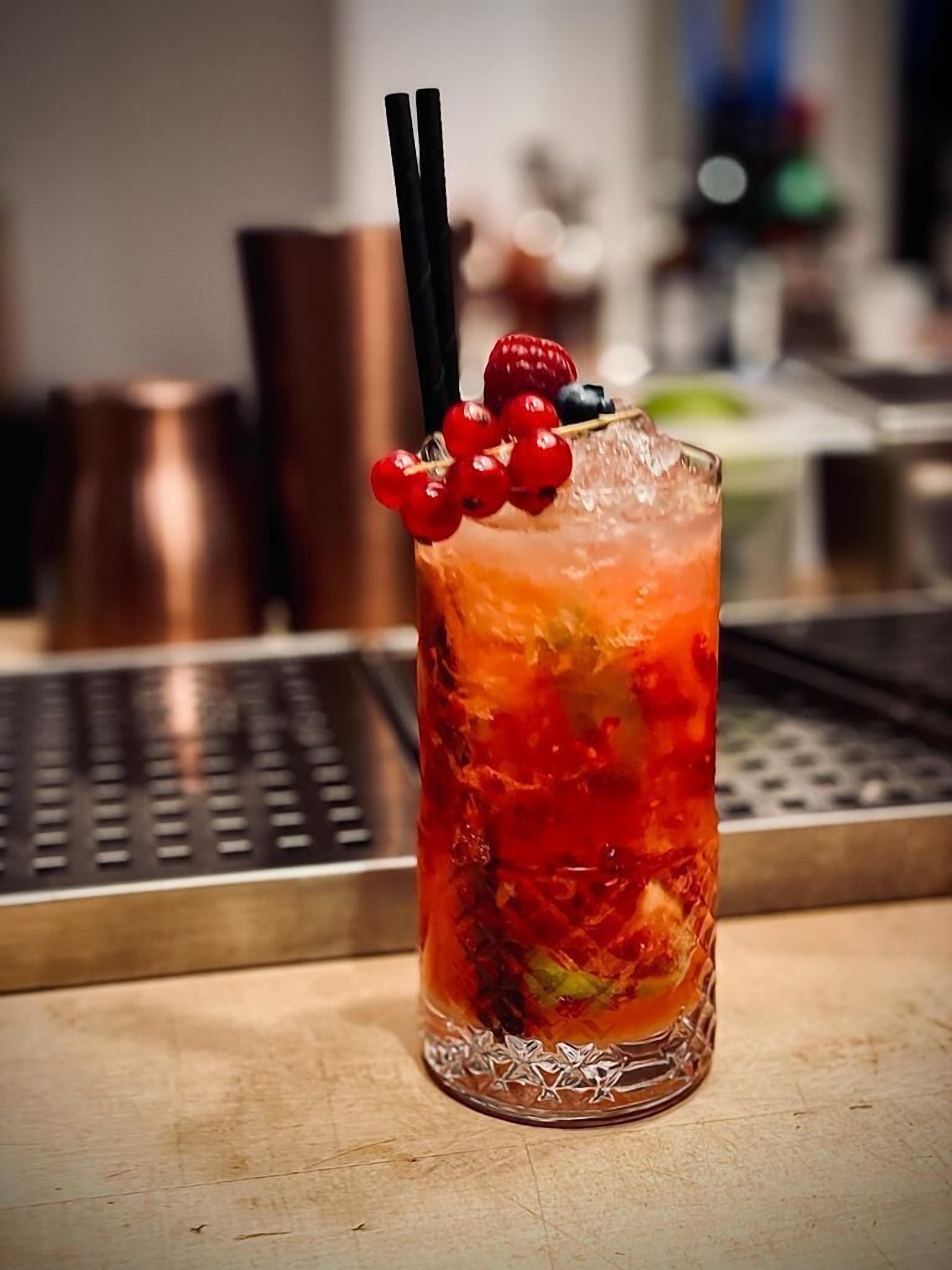 Ein Cocktailglas mit einem roten Getränk, verziert mit Beeren und einem Strohhalm, auf einer Bar.