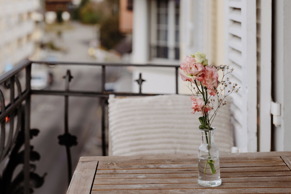 A wooden table with a vase of flowers on a balcony overlooking a residential area.