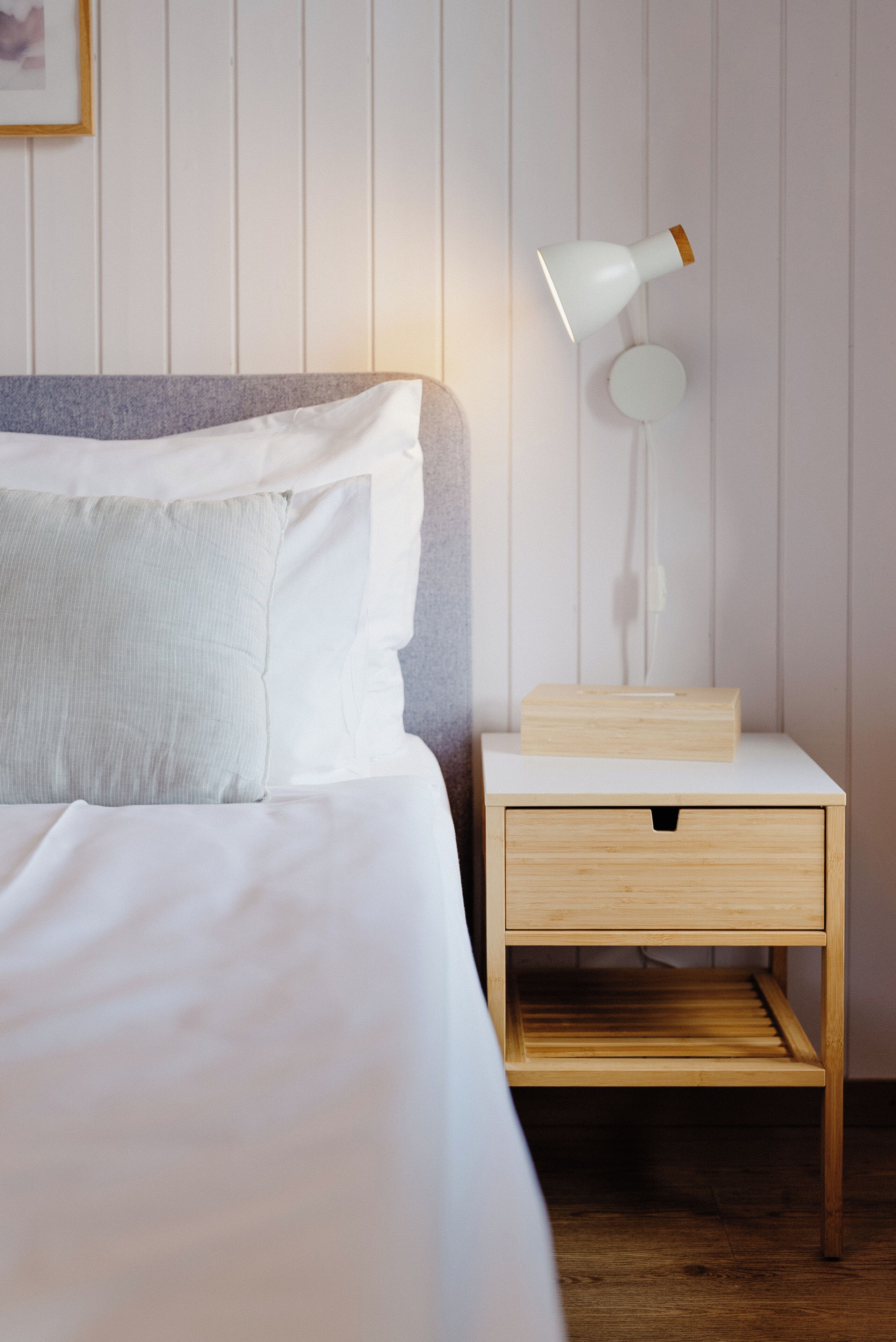 Una cama bien hecha con ropa blanca, una mesita de noche con lámpara y una mesita auxiliar de madera.