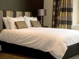 Una cama bien hecha con sábanas blancas y varios almohadones, una lámpara de noche y una cortina estampada.