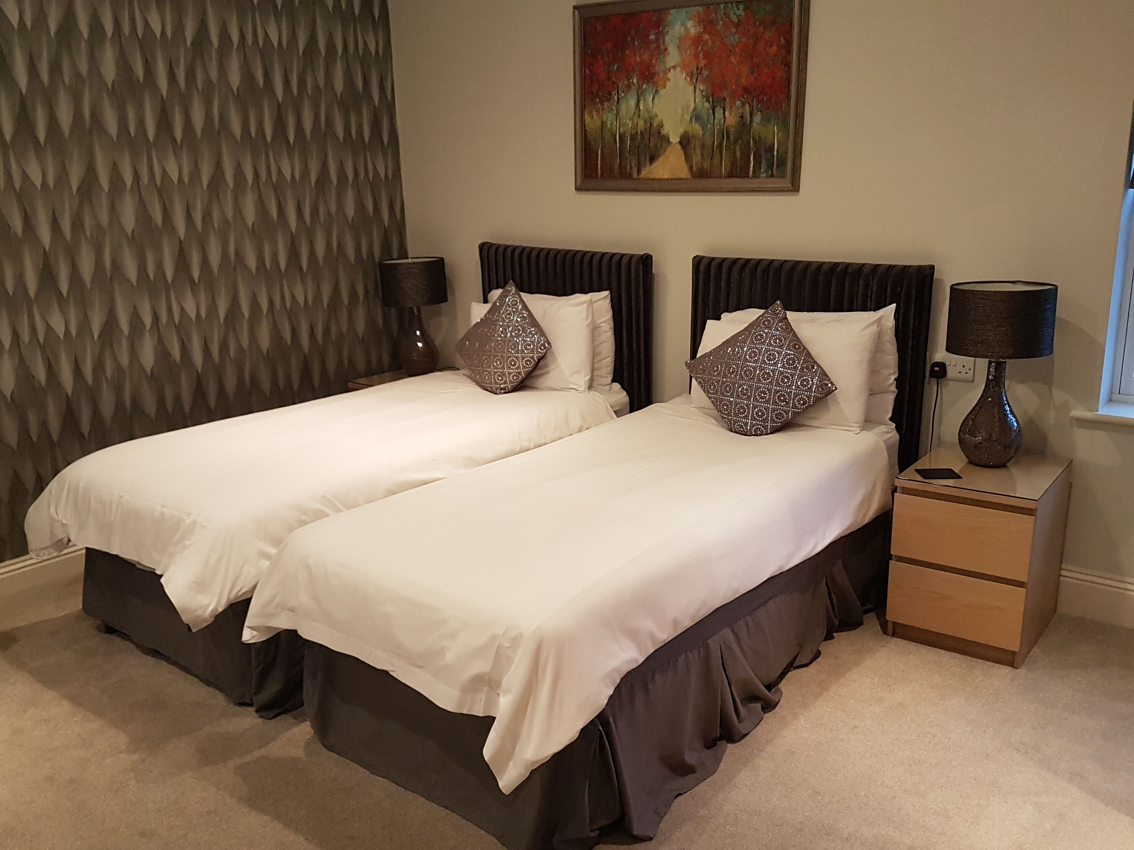 A double bed with a dark headboard and a decorative pillow, a bedside table with a lamp, and a painting on the wall.