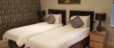 Una cama doble con cabecera oscura y un cojín decorativo, una mesita de noche con lámpara, y un cuadro en la pared.