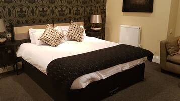 Una cama bien hecha con colcha blanco y negro, dos almohadas decorativas, una mesita de noche con una lámpara, un radiador y una pared con papel tapiz estampado.