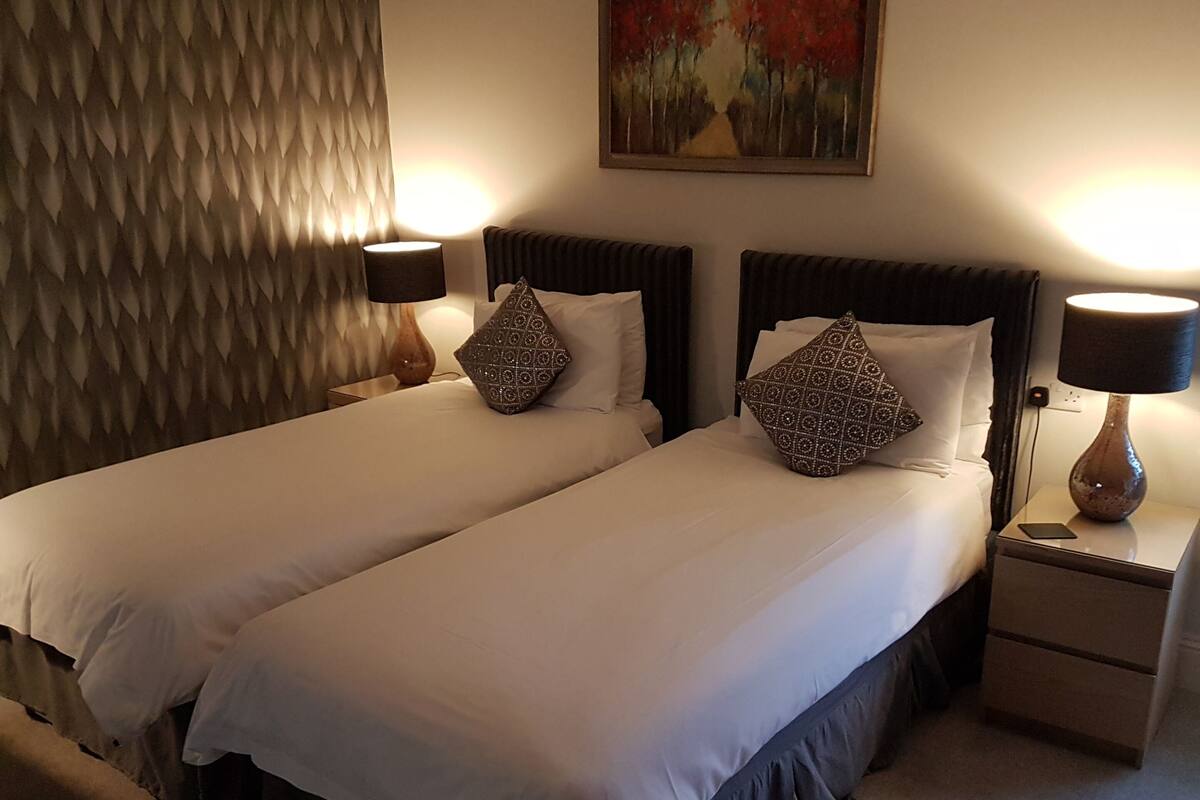 Hotellihuone, jossa on kaksi sänkyä, ja jokaisessa sängyssä on tyyny, lamppu ja seinällä taulu.