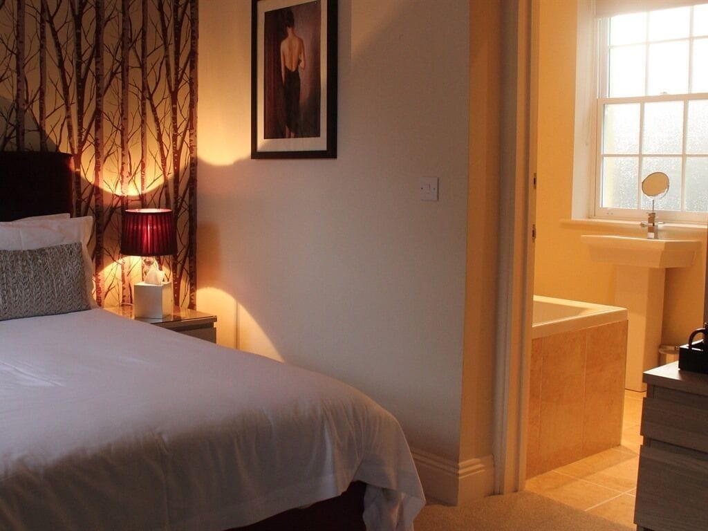 Un dormitorio con cama, mesita de noche, lámpara, un cuadro en la pared, ventana y baño con lavabo y espejo.