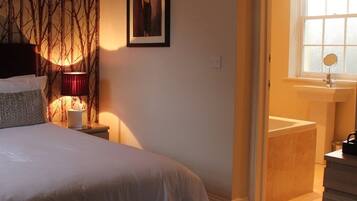 Un dormitorio con cama, mesita de noche, lámpara, un cuadro en la pared, ventana y baño con lavabo y espejo.