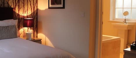 Ein Schlafzimmer mit Bett, Nachttisch, Lampe, einem gerahmten Bild an der Wand, einem Fenster und einem Badezimmer mit Waschbecken und Spiegel.