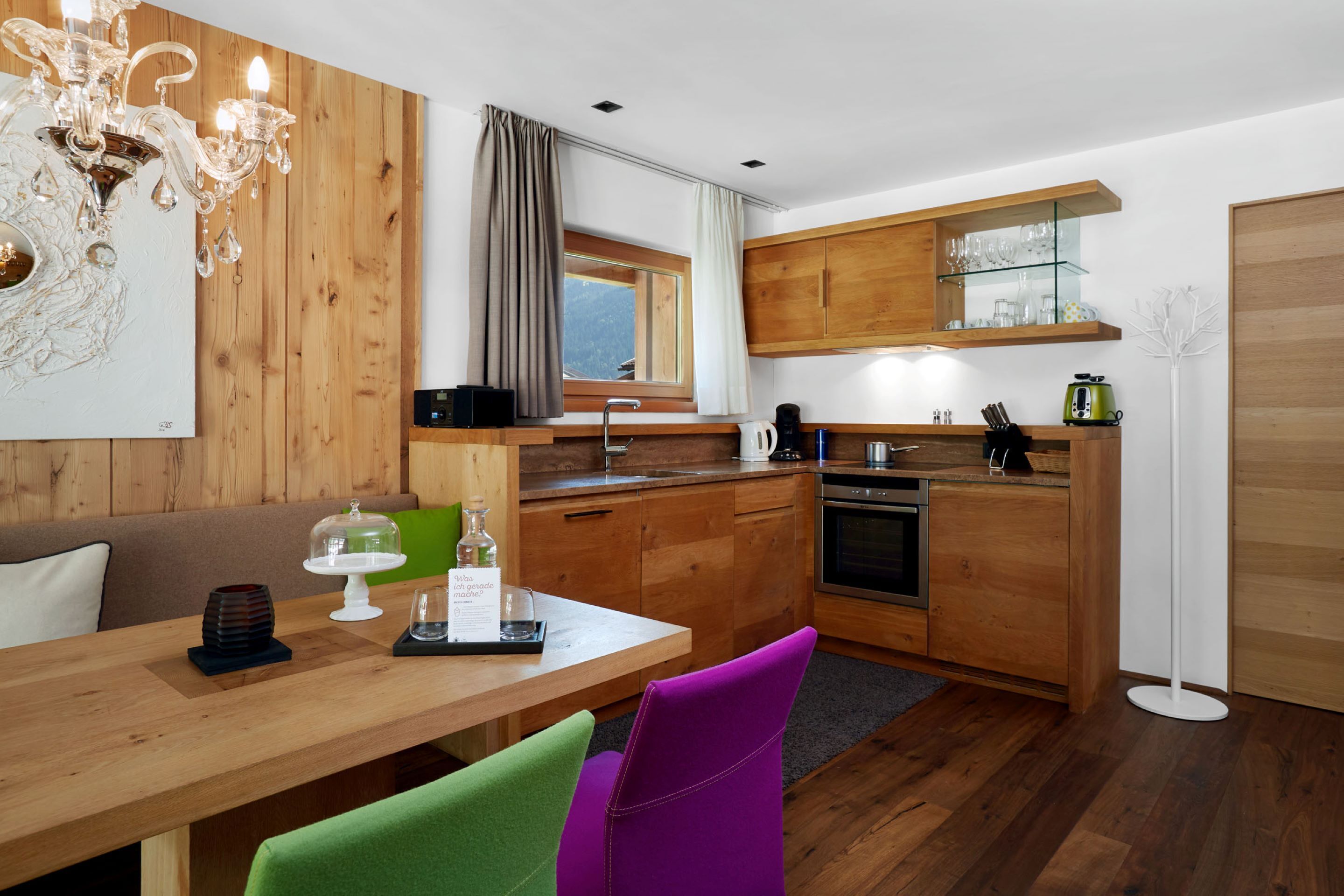A modern kitchen with wooden cabinets, a dining table with chairs, a chandelier, and a window with curtains.