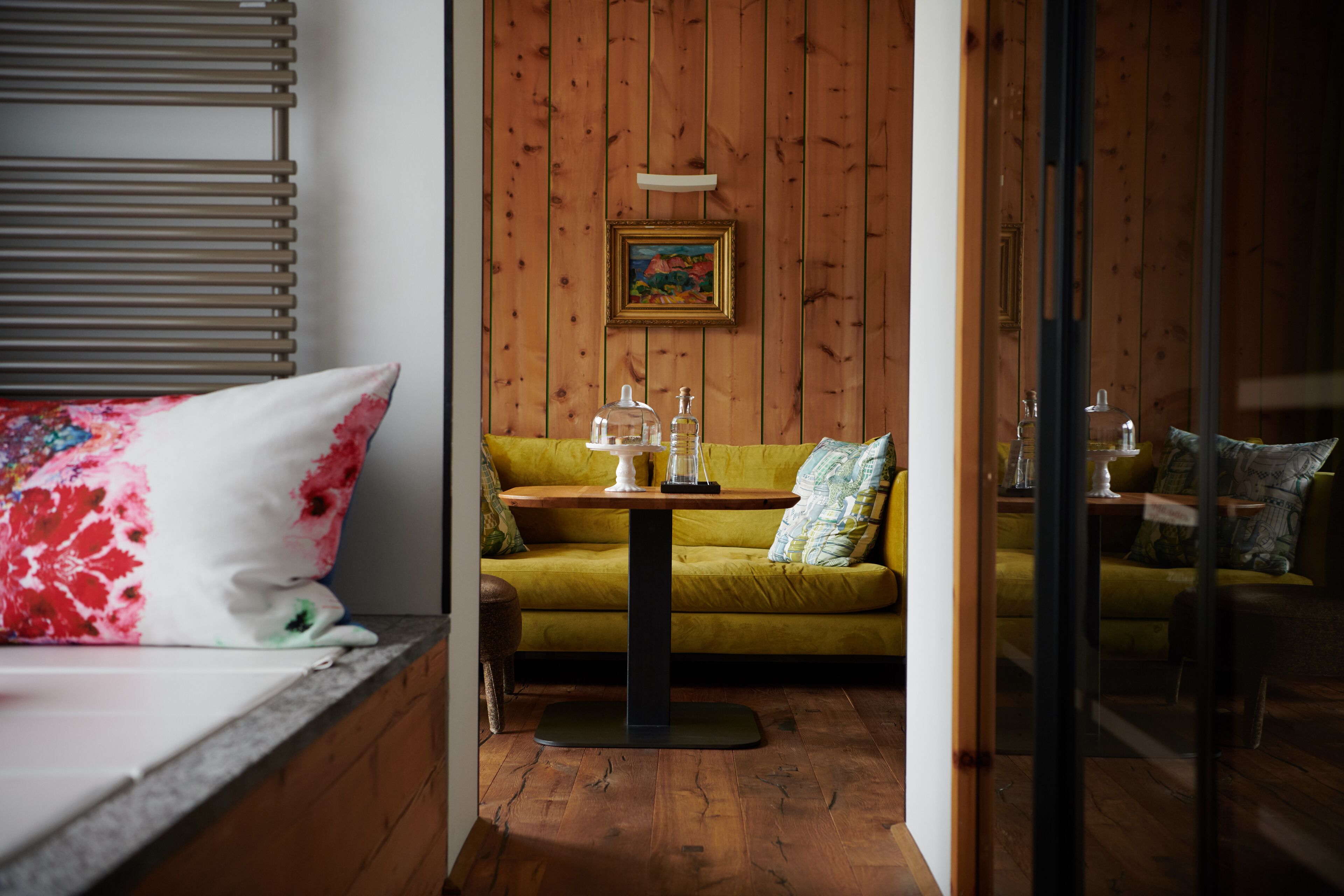 Een kamer met een bed, een gele bank, een klein rond tafeltje met een lamp en een houten wand met een lijst eromheen.