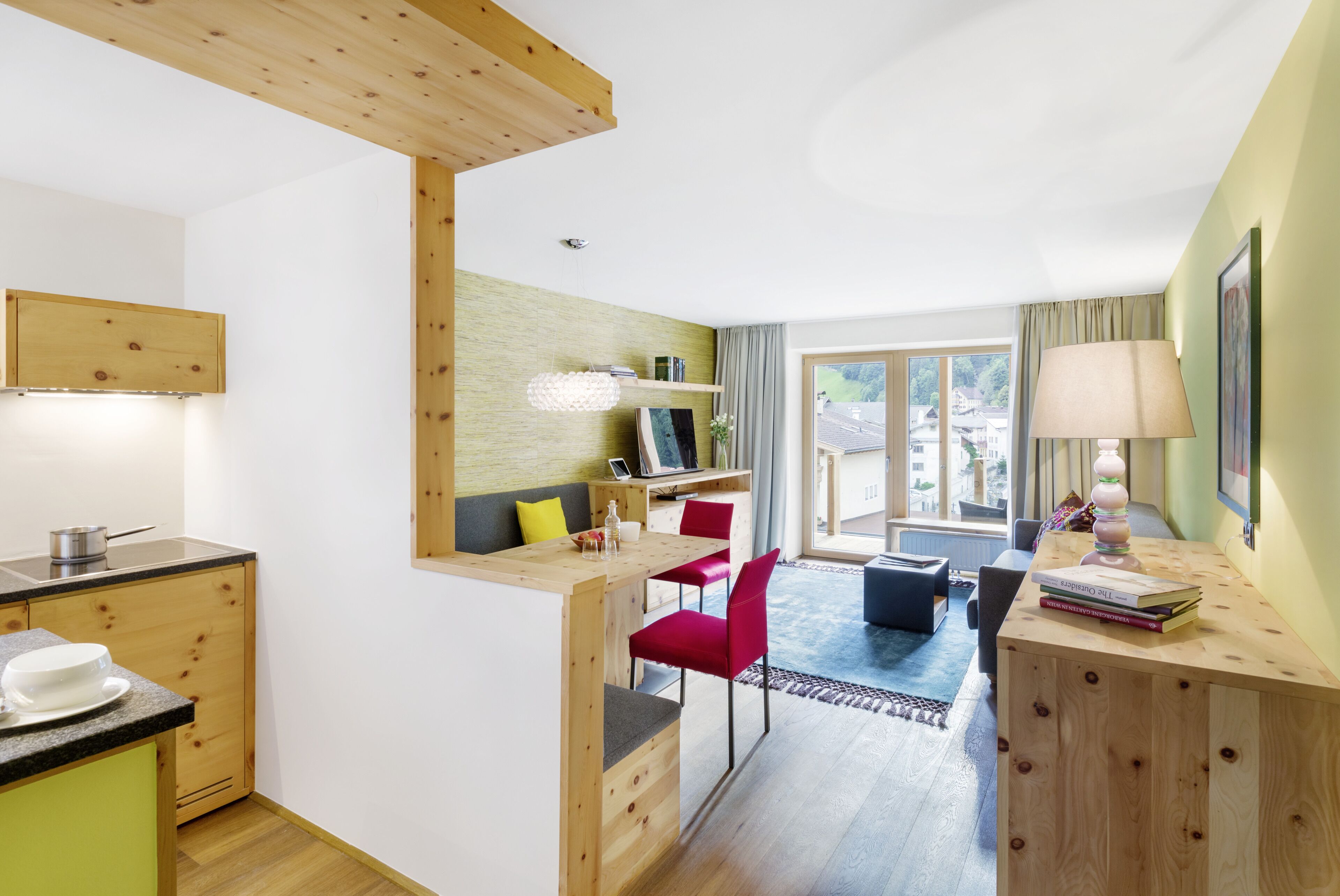 A modern kitchen with wooden cabinets, a dining area with red chairs, and a view of the outdoors through large windows.