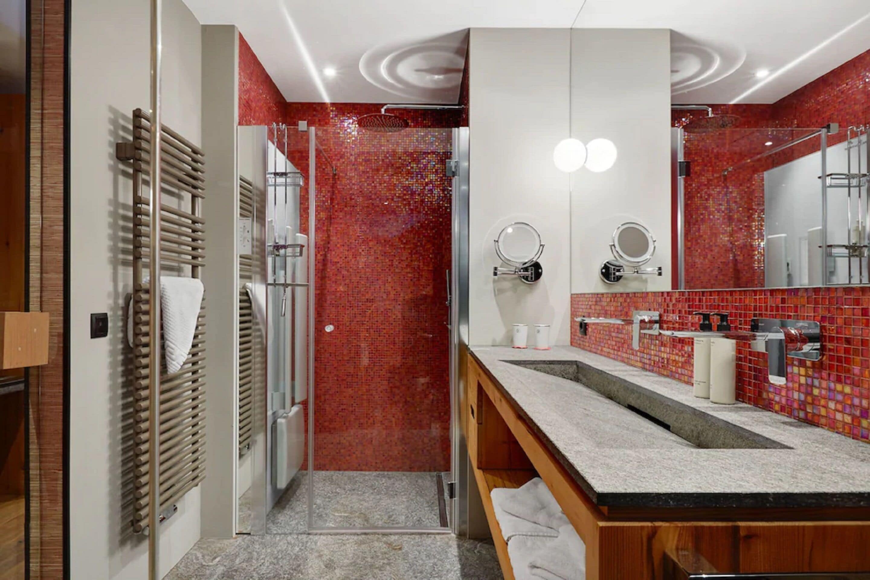 A modern bathroom with a glass shower, a wooden vanity, and a red mosaic tile wall.
