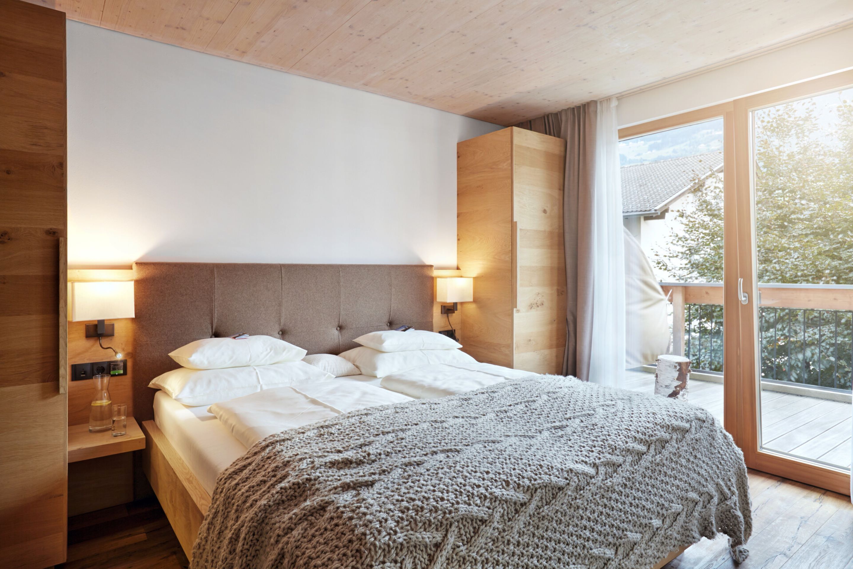 A bedroom with a large bed, wooden headboard, bedside lamps, and a view of the outdoors through a sliding glass door.