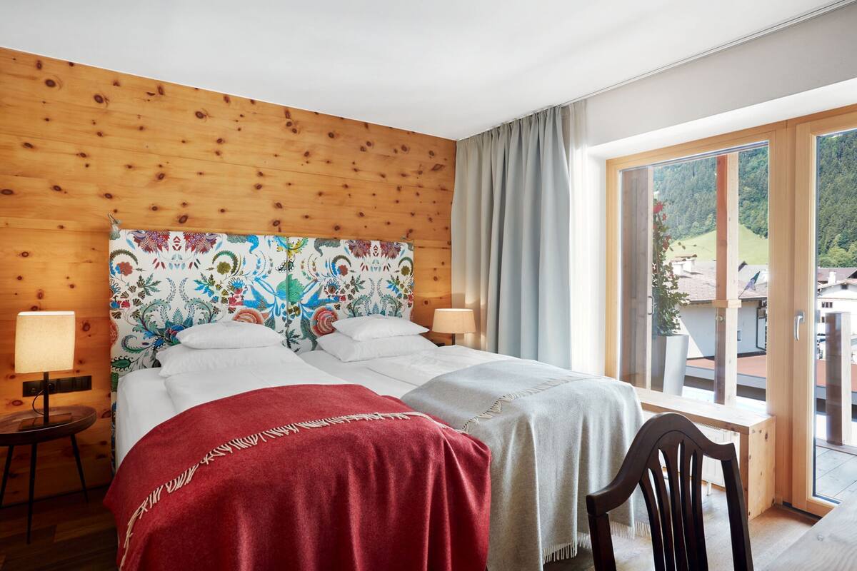 Une chambre à coucher avec une tête de lit en bois, un grand lit recouvert de linge de lit blanc, deux tables de chevet avec des lampes, et une vue sur un paysage montagneux par une grande fenêtre.