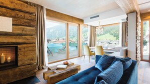 A modern living room with a large blue sofa, wooden walls, a fireplace, and a view of mountains and a pool. - DasPosthotel – Boutique Hotel (Zell am Ziller)